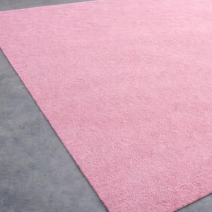 May include: A large, rectangular, solid pink rug. The rug has a soft, textured appearance and is placed on a gray concrete floor. The rug fills most of the frame, with a small portion of the floor visible in the corner.