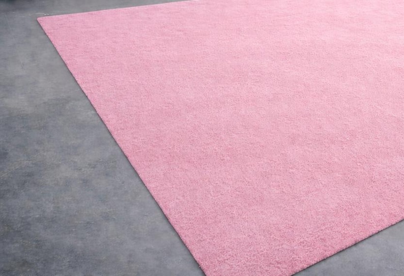 May include: A large, rectangular, solid pink rug. The rug has a soft, textured appearance and is placed on a gray concrete floor. The rug fills most of the frame, with a small portion of the floor visible in the corner.