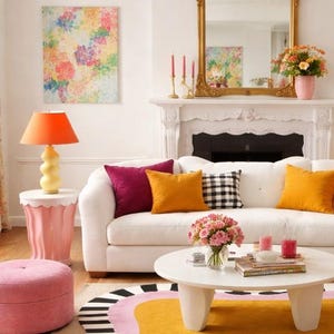 May include: A bright living room scene with a white sofa adorned with colorful pillows. A round coffee table holds flowers and candles. An orange lamp sits on a pink side table. A colorful rug and floral artwork complete the vibrant decor.