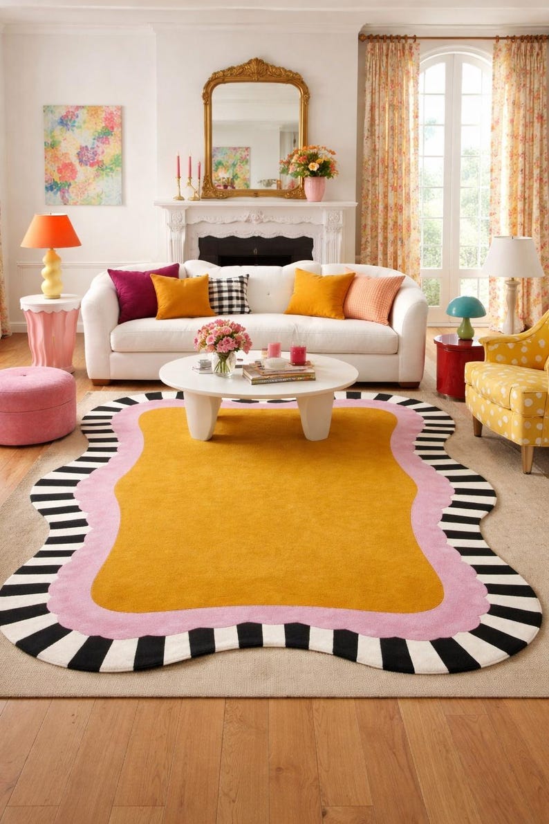May include: A living room scene featuring a large, mustard-yellow rug with a wavy pink border and black and white striped edges. The room includes a white sofa with colorful pillows, a gold mirror, and various lamps.