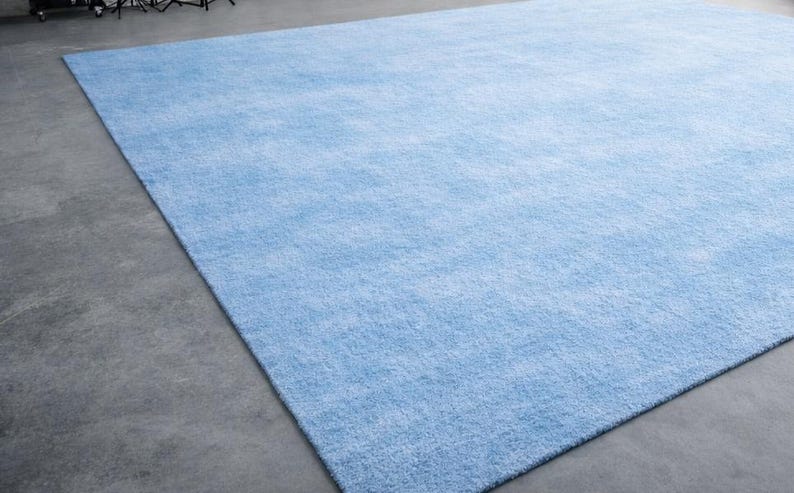 May include: A large, rectangular, light blue rug. The rug has a soft, textured appearance and is placed on a gray concrete floor. The rug's color is a solid, pale blue, and it fills most of the frame.