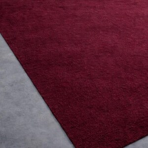 May include: A rectangular, burgundy-colored rug with a soft, textured surface. The rug is placed on a gray concrete floor, creating a contrast in color and texture. The rug's deep red hue adds warmth to the space.