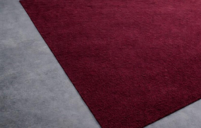 May include: A rectangular, burgundy-colored rug with a soft, textured surface. The rug is placed on a gray concrete floor, creating a contrast in color and texture. The rug's deep red hue adds warmth to the space.