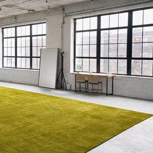 May include: A large, rectangular, lime green rug dominates the foreground of a bright, industrial-style room. The room features large, black-framed windows, white brick walls, and a concrete floor. A wooden table with two chairs and a large mirror are also present.