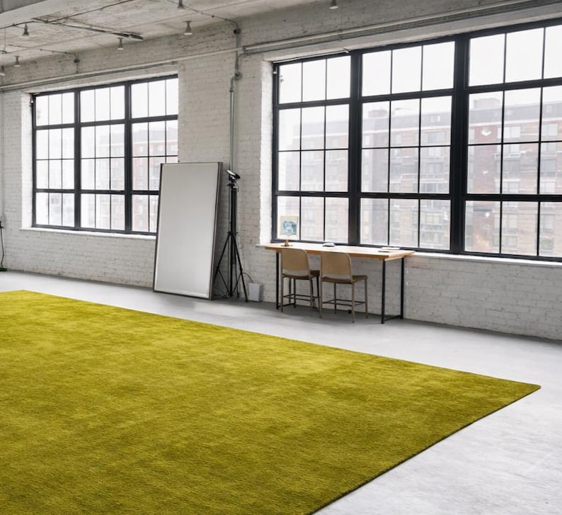 May include: A large, rectangular, lime green rug dominates the foreground of a bright, industrial-style room. The room features large, black-framed windows, white brick walls, and a concrete floor. A wooden table with two chairs and a large mirror are also present.