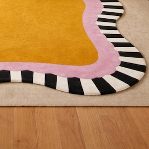 May include: A mustard yellow rug with a wavy edge, bordered by a light pink trim and a black and white striped pattern. The rug is placed on a neutral-colored carpet, with a wooden floor visible below.