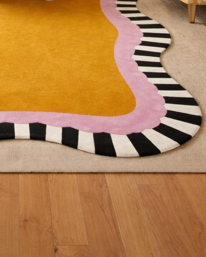 May include: A mustard yellow rug with a wavy edge, bordered by a light pink trim and a black and white striped pattern. The rug is placed on a neutral-colored carpet, with a wooden floor visible below.