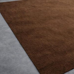 May include: A rectangular, brown rug with a soft, textured surface. The rug is placed on a gray concrete floor, with a portion of the floor visible in the image. The rug's color is a rich, warm brown.