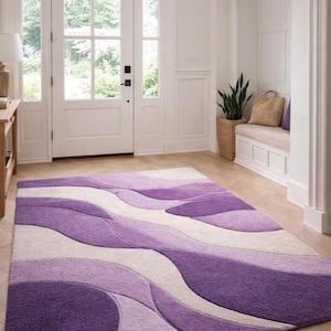 May include: A rectangular area rug with a wavy pattern in shades of purple and off-white. The rug is placed on a wooden floor in a well-lit entryway. The rug's design adds a modern touch to the space.