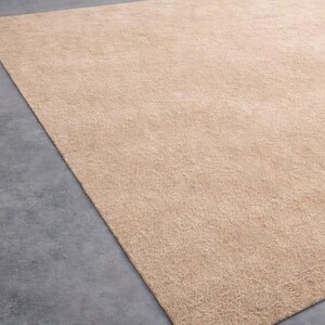 May include: A large, rectangular beige rug with a soft, textured surface. The rug is placed on a gray concrete floor, with a portion of the rug visible in the frame. The rug's color is a warm, neutral tone.