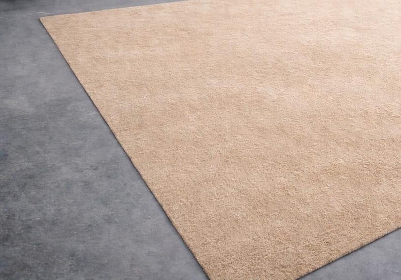 May include: A large, rectangular beige rug with a soft, textured surface. The rug is placed on a gray concrete floor, with a portion of the rug visible in the frame. The rug's color is a warm, neutral tone.