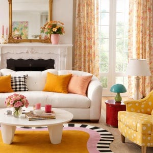 May include: A bright living room with a white sofa, yellow rug, and floral curtains. The sofa has orange and patterned pillows. A gold mirror hangs above a white fireplace. A yellow polka dot chair and a red side table add pops of color.