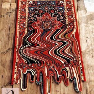 May include: A rectangular rug with a traditional Persian design, featuring a red border and a central pattern in black, blue, and red. The bottom portion of the rug has a melting effect, with vertical stripes in various colors. The rug is placed on a wooden surface.