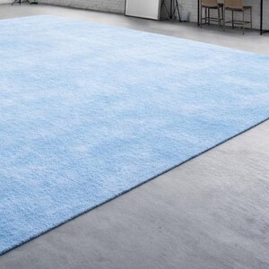 May include: A large, rectangular, light blue rug with a soft, textured appearance. The rug fills most of the frame, with a concrete floor visible in the foreground. The rug's color is a solid, pale blue, and it appears to be made of a plush material.