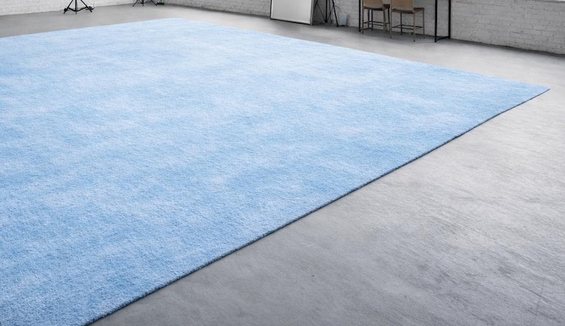 May include: A large, rectangular, light blue rug with a soft, textured appearance. The rug fills most of the frame, with a concrete floor visible in the foreground. The rug's color is a solid, pale blue, and it appears to be made of a plush material.