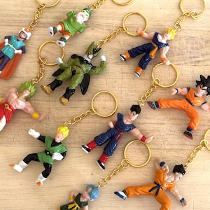 May include: A collection of colorful anime character keychains with gold-colored chains and rings. The figurines are in various action poses and feature characters in shades of green, orange, blue, and yellow. The keychains are arranged on a light-colored wooden surface.