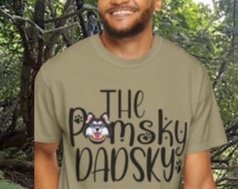 May include: A light olive green t-shirt with the text "The Pomsky DADSKY" in black script and a Pomsky dog graphic. A person is wearing the shirt and smiling. A Pomsky puppy is walking on a path in the background.