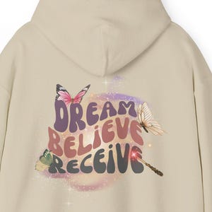 May include: Beige hoodie with the words "DREAM BELIEVE RECEIVE" in a colorful, whimsical font. The design includes butterfly illustrations. The hoodie has a hood and long sleeves.