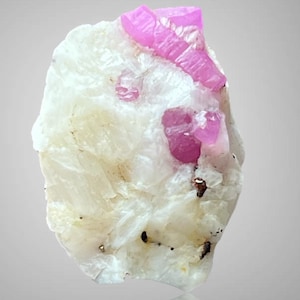 May include: A natural mineral specimen featuring a white matrix with embedded pink ruby crystals. The ruby crystals are a vibrant pink color and have a crystalline structure. The specimen is displayed against a light gray background.