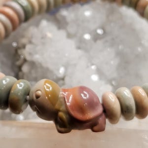 May include: A beaded bracelet featuring a turtle charm. The bracelet is made of small, round, multi-colored beads in shades of beige, pink, and green. The turtle charm is carved from a similar stone, with a detailed shell and head.