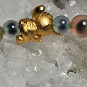 May include: A gold-coloured charm shaped like a small animal is the focal point of this bracelet. The bracelet features a series of colourful, round beads in shades of green, grey, and pink. The bracelet is displayed on a white, crystalline surface.