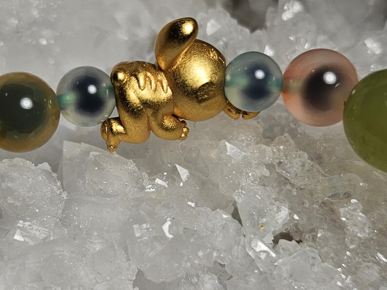 May include: A gold-coloured charm shaped like a small animal is the focal point of this bracelet. The bracelet features a series of colourful, round beads in shades of green, grey, and pink. The bracelet is displayed on a white, crystalline surface.