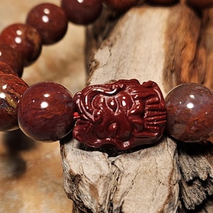 May include: A close-up of a beaded bracelet featuring large, round, reddish-brown beads. The bracelet includes a carved, dark red centerpiece. The beads have a polished finish, and the bracelet is displayed on a piece of wood.