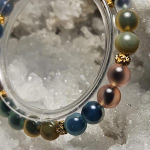 May include: A multi-coloured beaded bracelet with round beads in shades of blue, green, pink, and gold. The bracelet has gold-coloured spacers. The bracelet is displayed on a white, textured surface. The beads are approximately 1 cm in diameter.