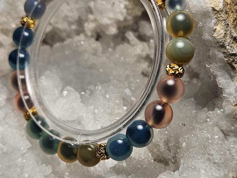 May include: A multi-coloured beaded bracelet with round beads in shades of blue, green, pink, and gold. The bracelet has gold-coloured spacers. The bracelet is displayed on a white, textured surface. The beads are approximately 1 cm in diameter.