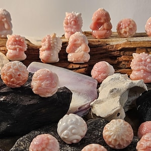 May include: A collection of pink and white carved shell figurines, some with intricate designs, arranged on a wooden surface and rocks. The figurines are small and detailed, with various shapes and sizes.
