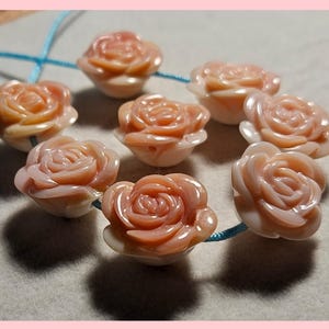 May include: A collection of peach-colored rose-shaped beads strung on a blue cord. The beads are intricately carved, showcasing the delicate petals of the roses. The beads are arranged in a circular pattern, perfect for jewelry making.