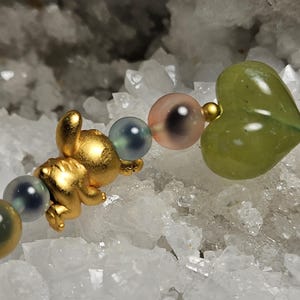 May include: A gold charm bracelet with a small bear, a green heart-shaped bead, and iridescent round beads. The bracelet is set against a background of white crystals, creating a contrast of textures and colours.