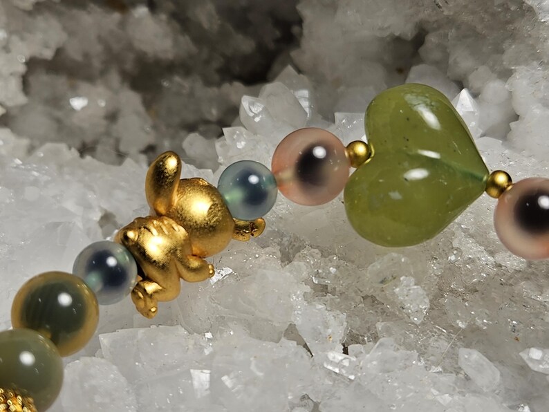 May include: A gold charm bracelet with a small bear, a green heart-shaped bead, and iridescent round beads. The bracelet is set against a background of white crystals, creating a contrast of textures and colours.