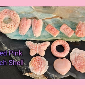 May include: A collection of carved pink conch shell charms, including a butterfly, a flower, a heart, a ring, and a dragonfly. The charms are displayed on a light blue and gray surface.