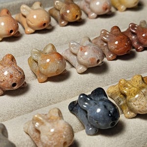 May include: A collection of small, carved stone animal figurines, resembling rabbits or similar creatures. The figurines are in various colors, including beige, brown, and gray, with a polished finish. Each has a small hole, possibly for stringing.