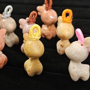 May include: A collection of carved stone deer figurines in various colors, including pink, beige, and yellow. Each figurine has a loop of cord at the top, in colors like orange, yellow, and pink. The deer are displayed on a black surface.