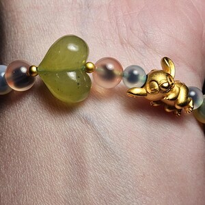 May include: A bracelet with a gold-coloured cartoon character charm, a green heart-shaped bead, and other beads in shades of blue, pink, and gold. The bracelet is worn on a wrist.