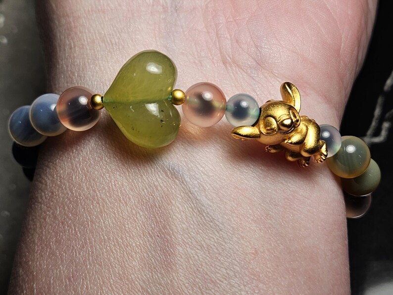 May include: A bracelet with a gold-coloured cartoon character charm, a green heart-shaped bead, and other beads in shades of blue, pink, and gold. The bracelet is worn on a wrist.