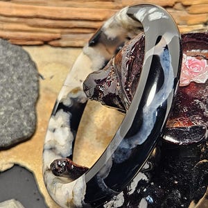 May include: A polished bangle bracelet featuring a smooth, rounded profile. The surface displays a high-contrast pattern of deep black sections interspersed with cloudy, translucent white and grey inclusions that resemble organic, swirling formations.