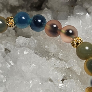 May include: A beaded bracelet with a variety of colourful, round gemstones in shades of blue, green, and pink. Small, gold-coloured decorative elements separate the beads. The bracelet is set against a backdrop of white, crystalline formations.