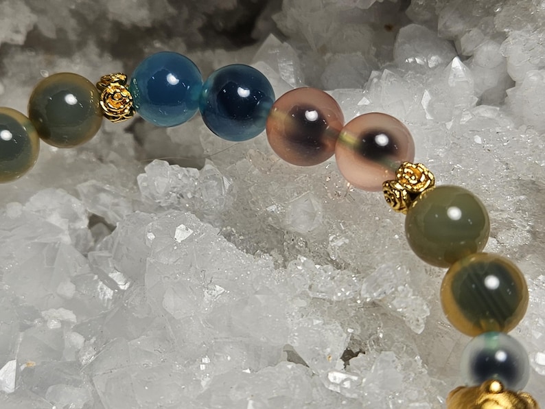 May include: A beaded bracelet with a variety of colourful, round gemstones in shades of blue, green, and pink. Small, gold-coloured decorative elements separate the beads. The bracelet is set against a backdrop of white, crystalline formations.