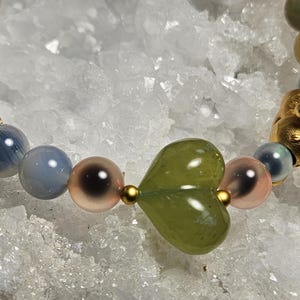 May include: A beaded bracelet with a gold pig charm, a green heart-shaped bead, and round beads in blue, pink, and brown. The bracelet is strung on a clear elastic cord. The background is white and crystalline.