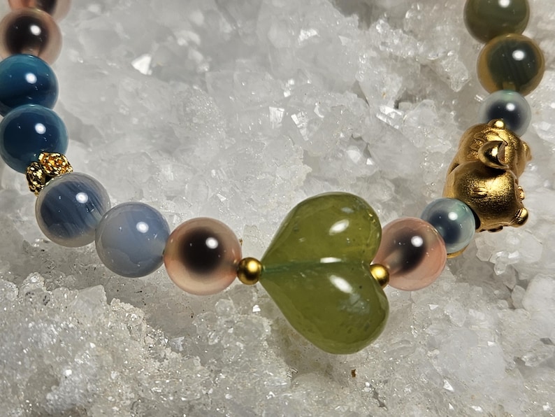 May include: A beaded bracelet with a gold pig charm, a green heart-shaped bead, and round beads in blue, pink, and brown. The bracelet is strung on a clear elastic cord. The background is white and crystalline.