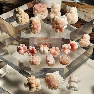 May include: A collection of 17 pink and white coral beads, each carved into a different animal or floral design. The beads are displayed on a tiered acrylic stand.
