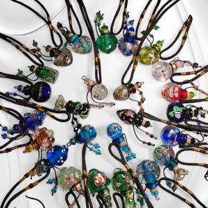 May include: A collection of colourful glass bottles with intricate designs, each hanging from a black cord with beads. The bottles are filled with different coloured liquids and have a variety of shapes and sizes.
