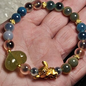 May include: A colourful beaded bracelet with blue, green, and pink gemstones. The bracelet has a gold-coloured rabbit charm and a heart-shaped stone. Gold-coloured spacers separate the beads. The bracelet is held in a hand.