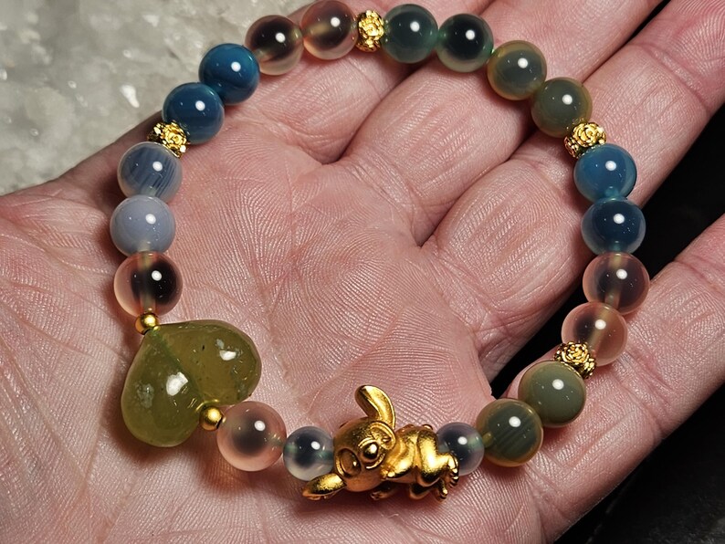 May include: A colourful beaded bracelet with blue, green, and pink gemstones. The bracelet has a gold-coloured rabbit charm and a heart-shaped stone. Gold-coloured spacers separate the beads. The bracelet is held in a hand.