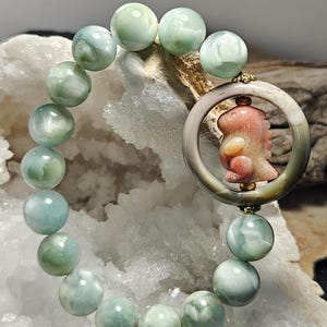 May include: A beaded bracelet featuring smooth, round, pale green and white marbled stone beads. The bracelet includes a circular pendant with a smooth outer rim and a carved, orange-toned creature figure positioned in the center of the ring.