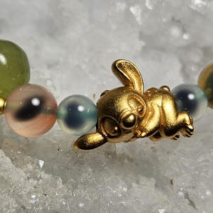 May include: A gold bunny charm bracelet featuring a mix of green, pink, and blue beads. The bunny is in a playful pose, adding a whimsical touch to the jewellery. The beads vary in size and colour.