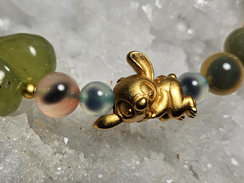 May include: A gold bunny charm bracelet featuring a mix of green, pink, and blue beads. The bunny is in a playful pose, adding a whimsical touch to the jewellery. The beads vary in size and colour.